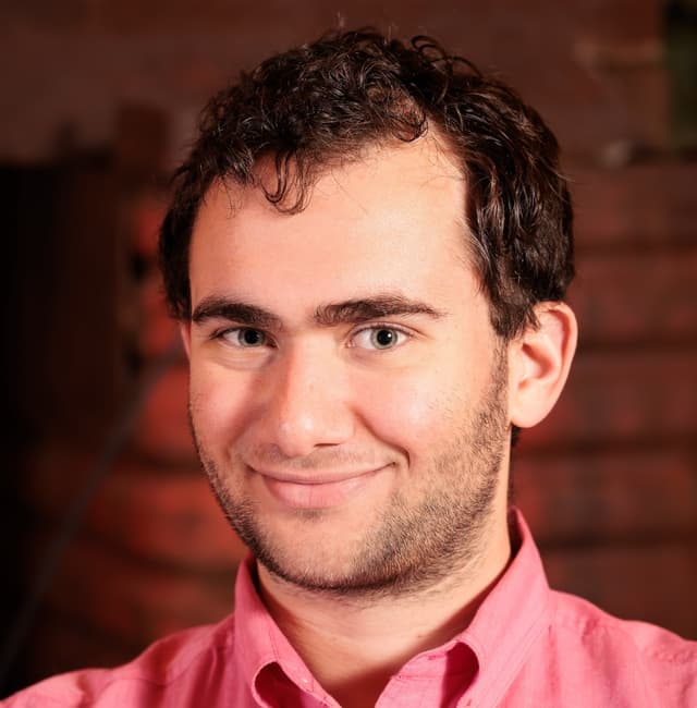 Shea Belsky wearing a red suit, black shirt, and black tie against a beige background.